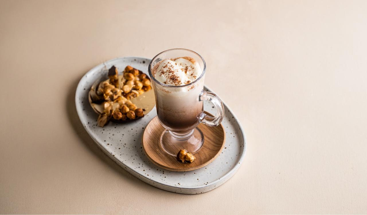 Glas mit Irish Coffee, bedeckt mit Schlagsahne und Kakaopulver, auf einem ovalen Teller mit Walnüssen