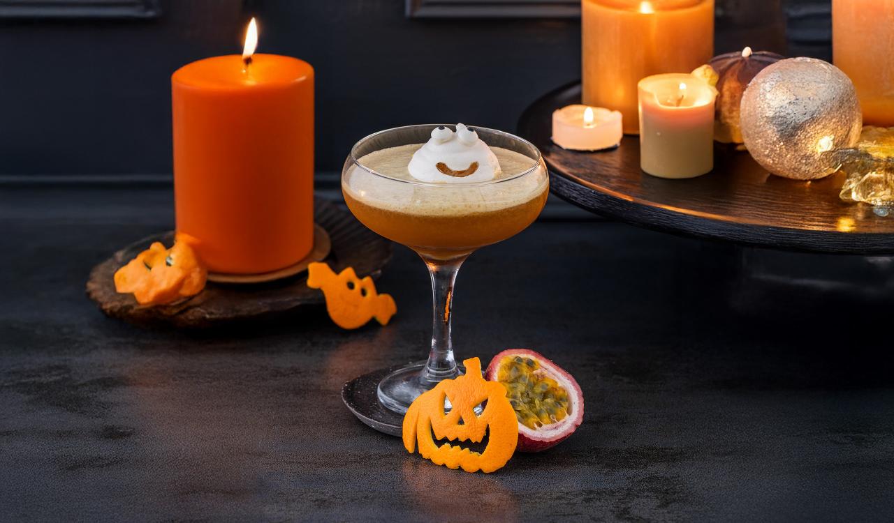 Cocktailglas mit Schaumhaube und lachendem Kürbis aus Orangenschale daneben, umgeben von brennenden Kerzen und einer halbierten Passionsfrucht auf dunklem Untergrund