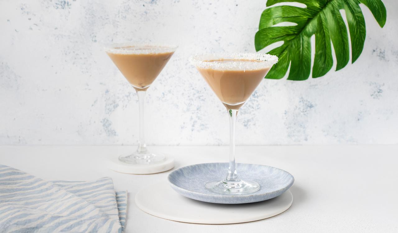 Zwei Martini-Gläser mit cremigem Kokos-Cocktail auf hellem Untergrund, eines auf einem grauen Teller, daneben ein grünes Blatt und ein gestreiftes Tuch