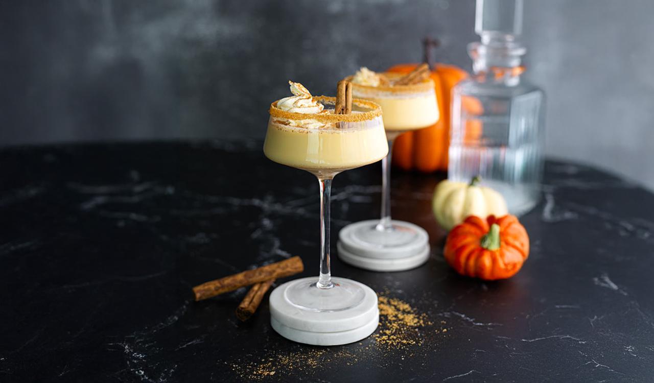 Zwei Cocktails mit Schlagsahne und Zimt auf Marmoruntersetzern vor Kürbissen und Glasflasche auf dunklem Untergrund
