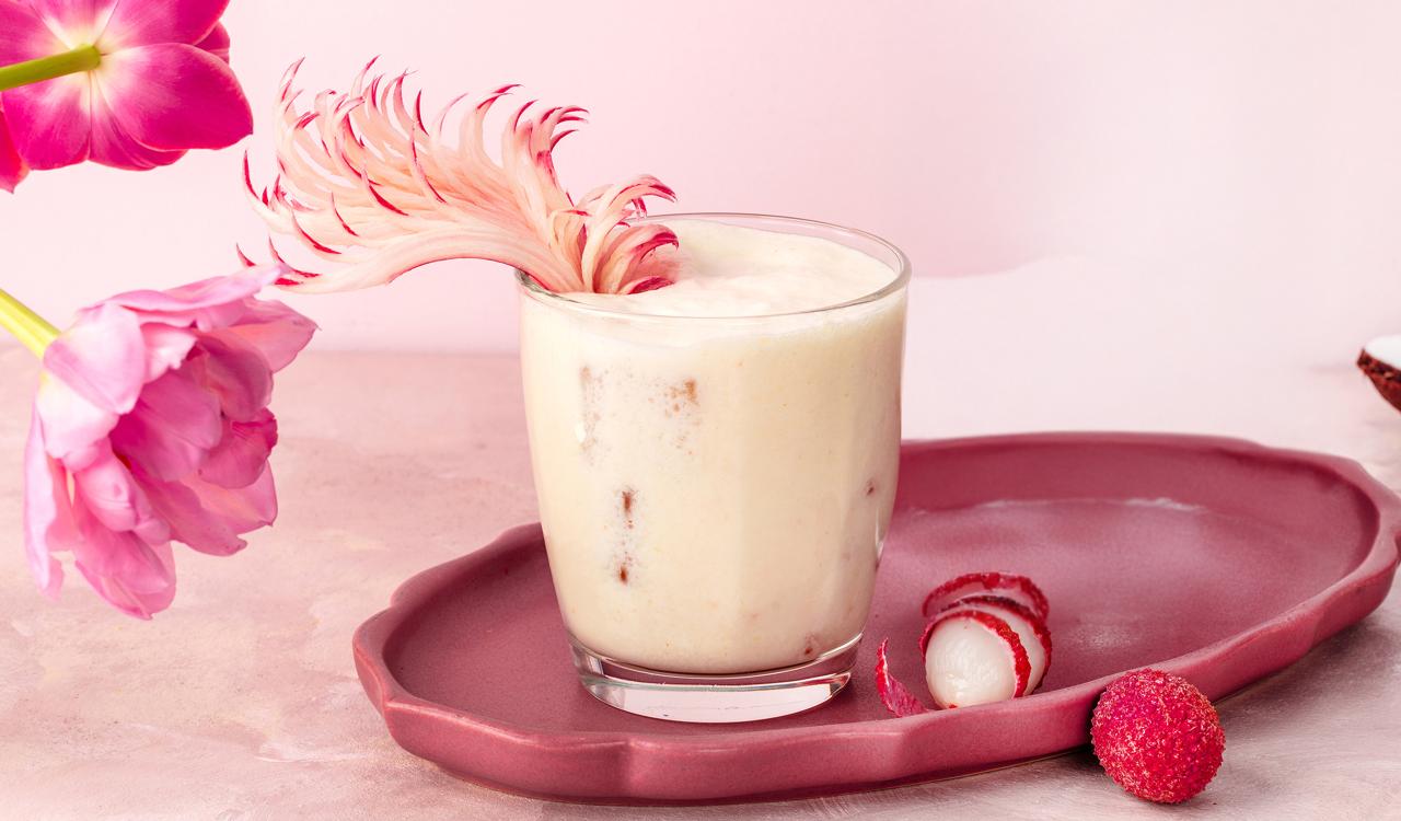 Glas mit cremigem Cocktail auf rosa Tablett neben zwei kleinen roten Kugeln und rosa Blume auf hellem Untergrund