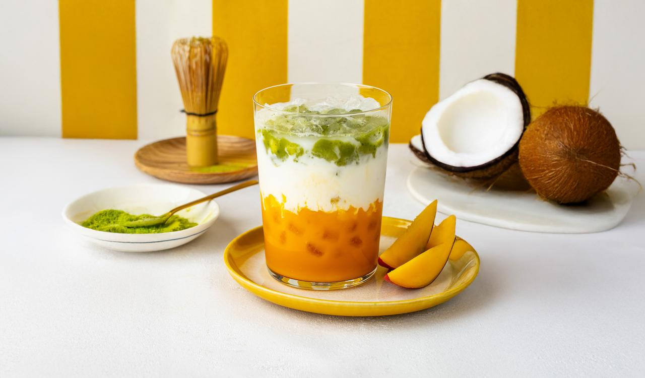 Glas mit geschichteter Mango-Matcha-Latte auf gelbem Teller mit Mangoscheiben daneben und halbierter Kokosnuss im Hintergrund