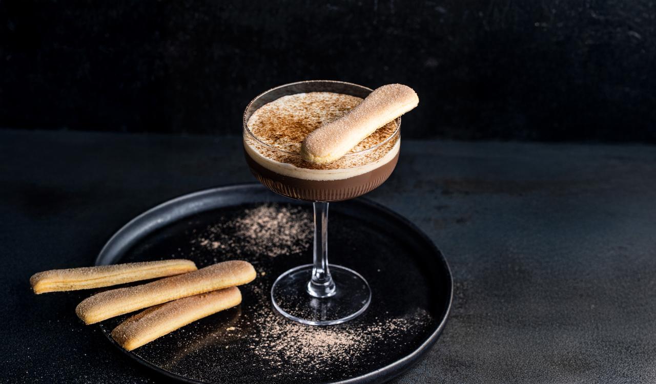 Cocktailglas mit Tiramisu-Martini und Löffelbiskuit auf schwarzem Teller vor dunklem Hintergrund