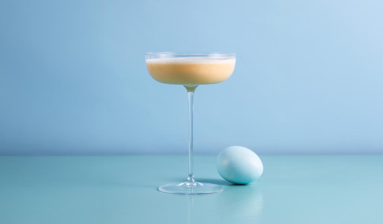 Cocktailglas mit cremigem hellgelbem Getränk neben einem weißen Ei auf hellblauem Untergrund vor hellblauem Hintergrund