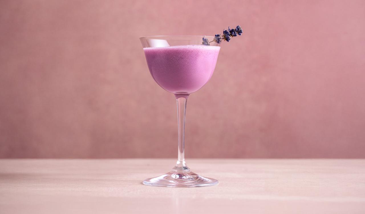 Cocktailglas mit cremigem, hellviolettem Getränk und Lavendelzweig als Garnitur vor einfarbigem Hintergrund