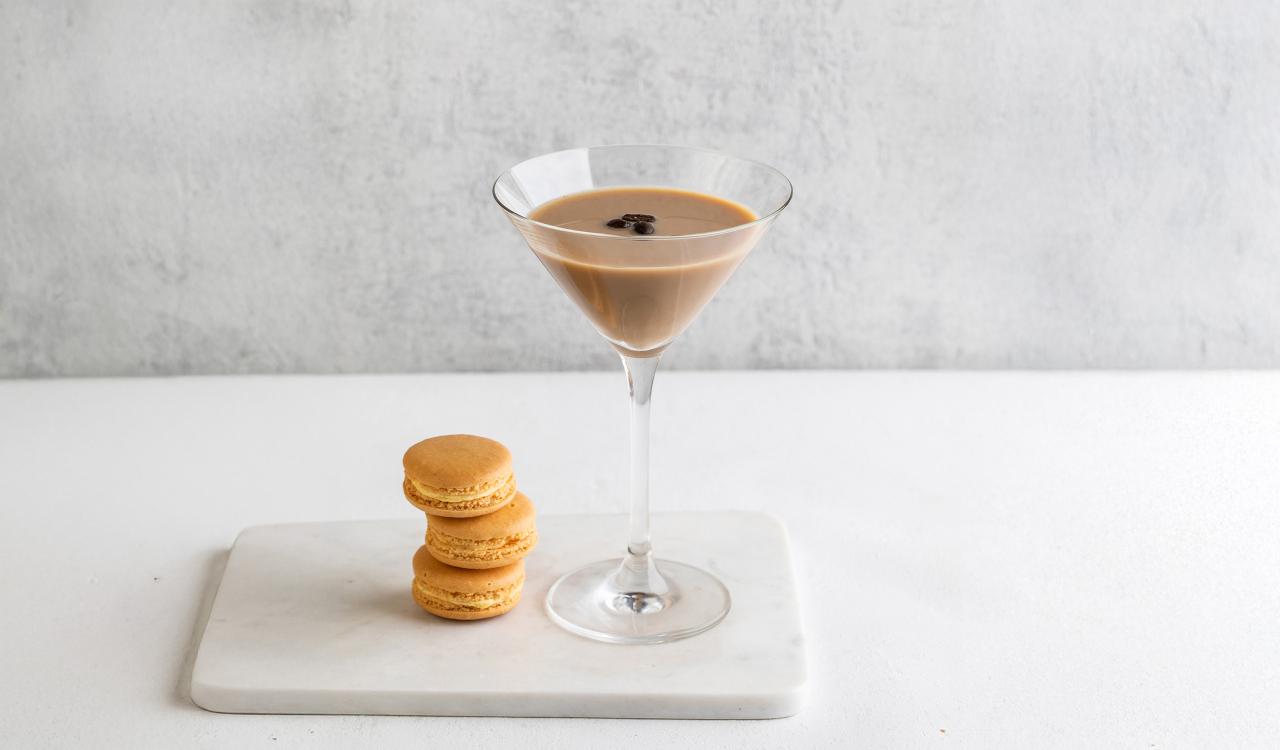 Martiniglas mit hellbraunem Espresso-Martini und drei Kaffeebohnen darauf neben zwei gestapelten hellbraunen Macarons auf weißem Marmorbrett vor hellgrauem Hintergrund