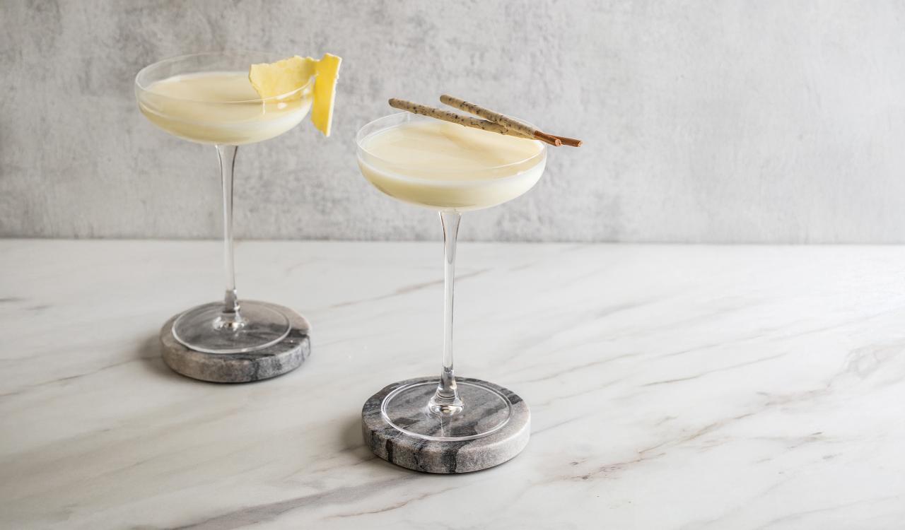 Zwei Cocktails in Martinigläsern mit weißer Schaumkrone auf Marmoruntersetzern, einer mit Zitronenscheibe, der andere mit zwei Zimtstangen garniert