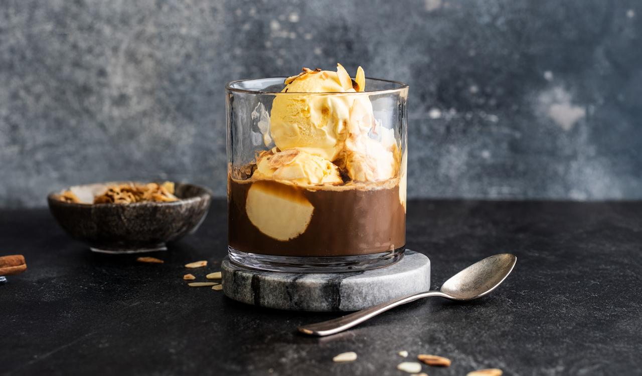 Glas mit Affogato bestehend aus Vanilleeis und Espresso auf grauem Marmoruntersetzer mit Löffel und Schale im Hintergrund