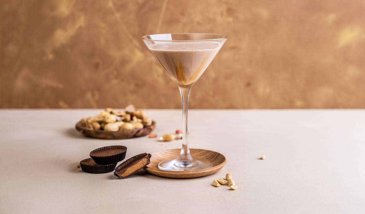Nahaufnahme eines Martini-Glases mit cremigem Getränk und Erdnussbutter, daneben Schalen mit Erdnussbutter-Pralinen und Erdnüssen auf hellem Untergrund