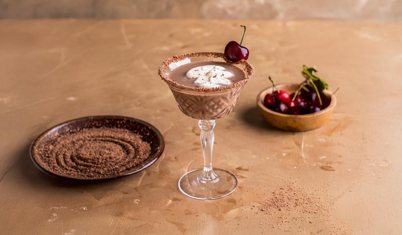 Cocktailglas mit Schokoladenmousse, Sahnehaube und Kirsche auf beigem Untergrund neben Schale mit Kirschen und Teller mit Kakaopulver