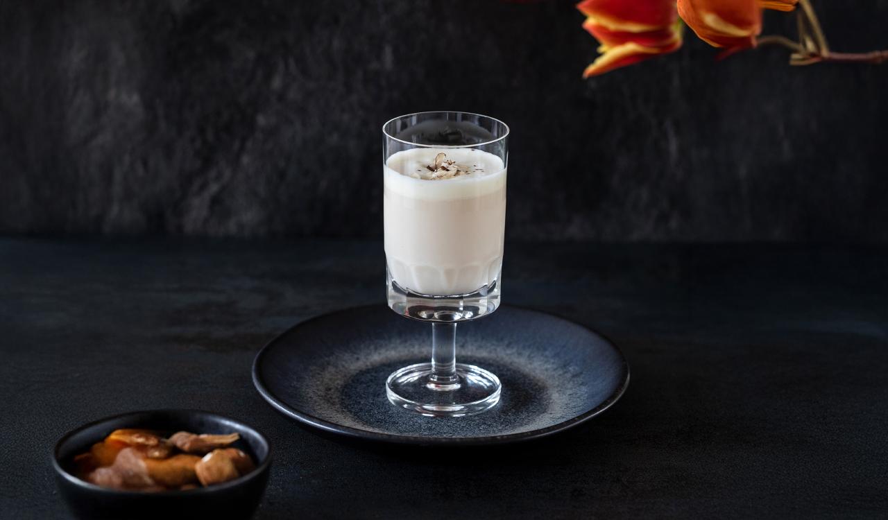 Glas mit weißem Getränk auf dunklem Teller vor dunklem Hintergrund und orangefarbenen Blüten im Hintergrund
