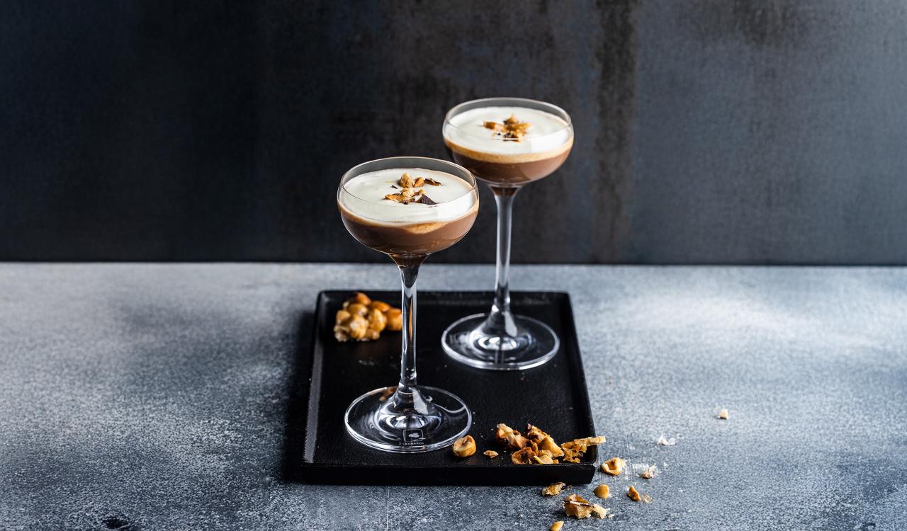 Zwei Cocktailgläser mit schaumiger Kaffee-Cocktail-Mischung auf schwarzem Tablett mit Kaffeebohnen und Kaffeebohnenstückchen auf grauem Untergrund
