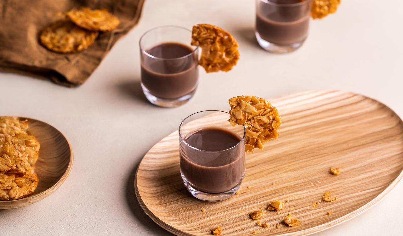 Drei Gläser mit braunem Cocktail, jedes mit einem Mandelkeks am Glasrand, auf hellem Untergrund und Holztablett