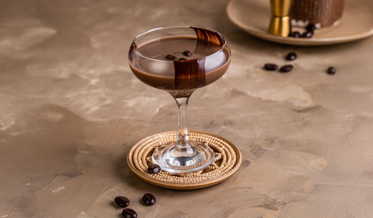 Cocktailglas mit dunkelbraunem Getränk, verziert mit Kaffeebohnen und Schokoladenstreifen, auf rundem Untersetzer