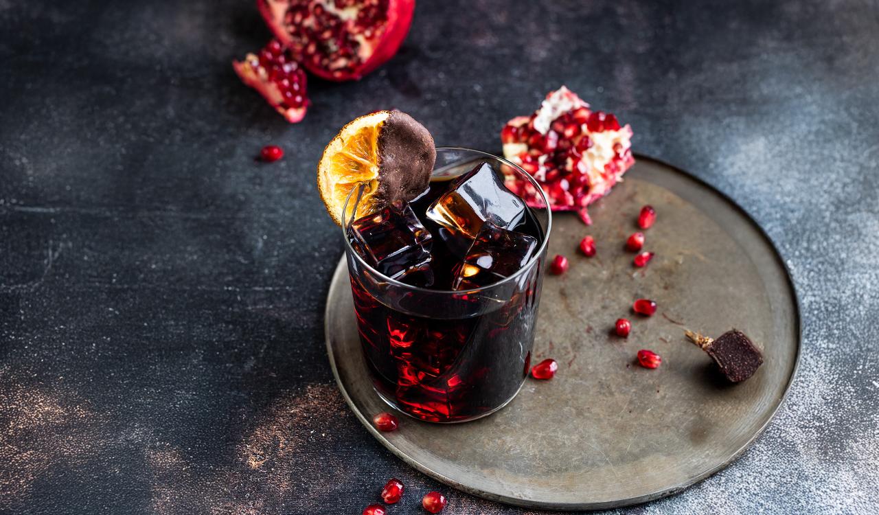 Glas mit dunklem Cocktail, Eiswürfeln und getrockneter Orangenscheibe auf grauem Teller mit Granatapfelkernen und aufgebrochenem Granatapfel im Hintergrund