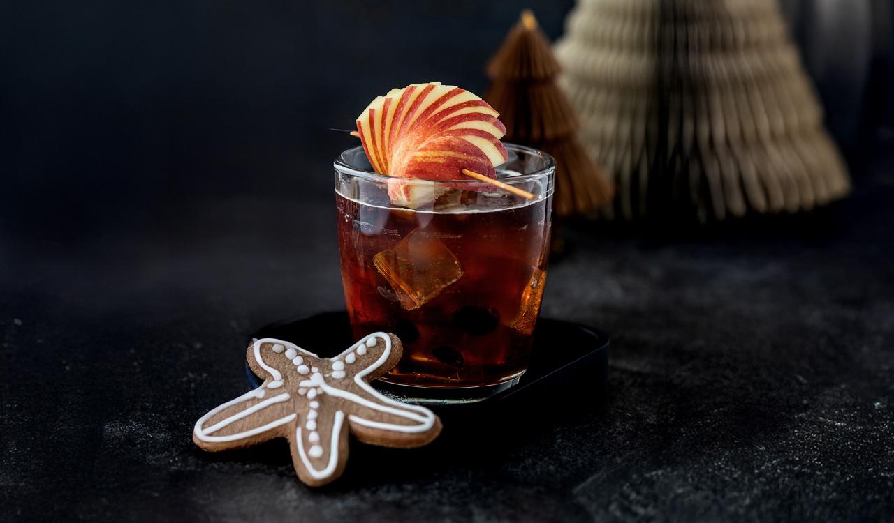 Glas mit dunklem Getränk, Eiswürfeln und Apfelscheiben als Garnitur neben sternförmigem Lebkuchen auf dunklem Untergrund