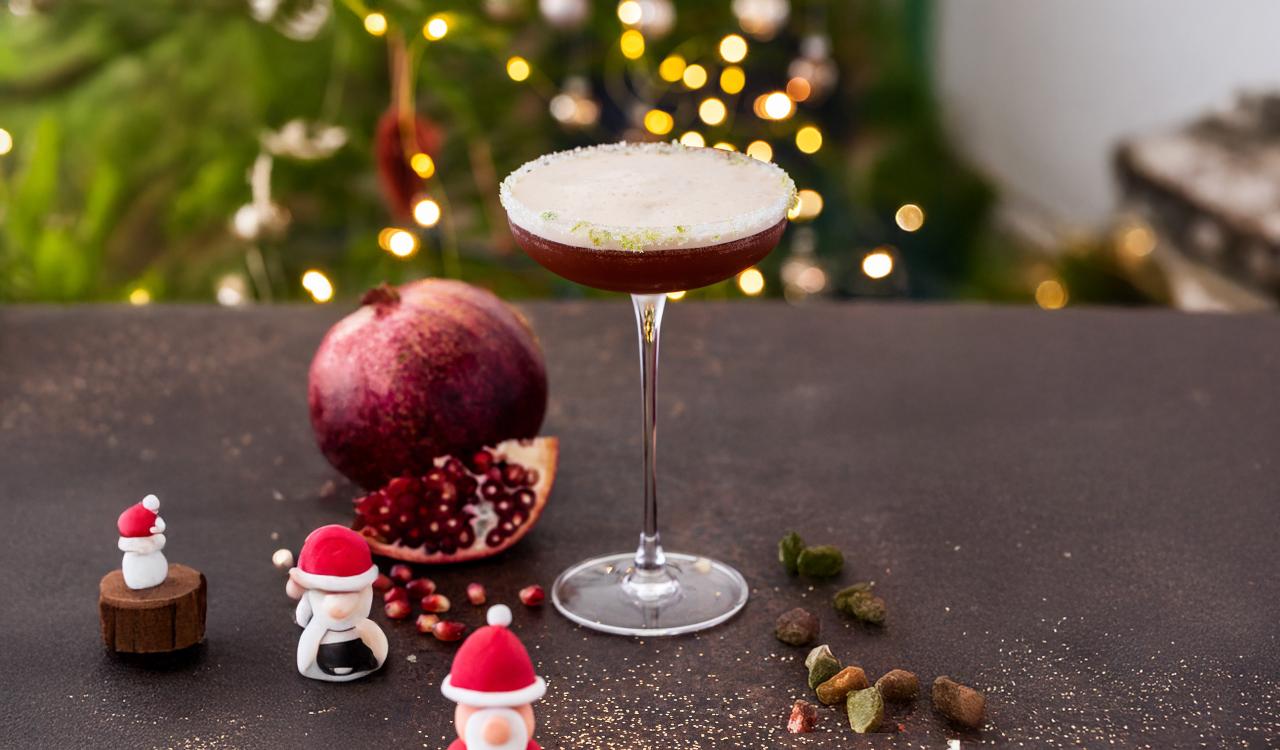 Cocktailglas mit schaumiger weißer Oberfläche auf dunklem Untergrund, im Hintergrund Granatapfel und kleine Figuren mit Weihnachtsmützen