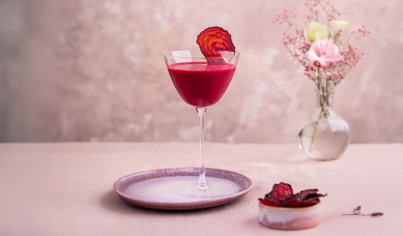 Cocktailglas mit dunkelrotem Getränk und getrockneter Fruchtscheibe auf einem Teller daneben ein kleines Glas mit weiteren getrockneten Fruchtscheiben und im Hintergrund eine Vase mit rosa Blumen