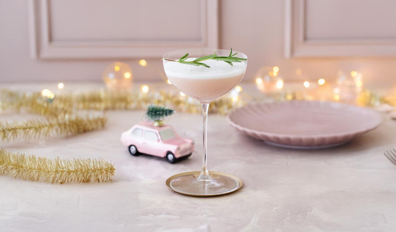A cocktail glass containing a white drink and sprigs of rosemary on a table with a pink plate and a small toy car with a Christmas tree on its roof