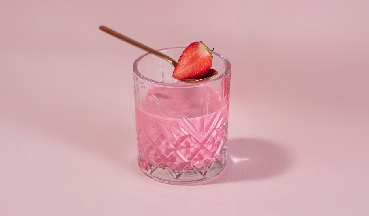 Kristallglas mit rosa Cocktail und halbierter Erdbeere auf Glasrand vor rosa Hintergrund