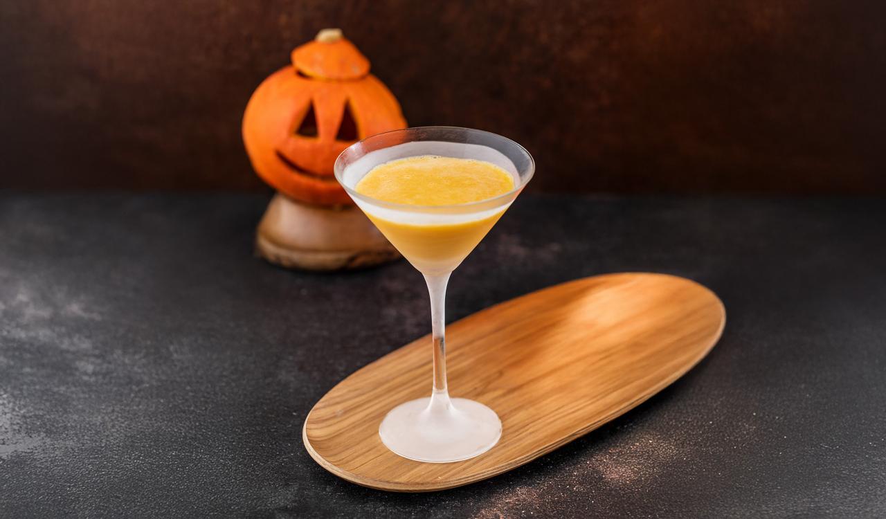 Cocktailglas mit gelb-orangefarbener Flüssigkeit auf einem länglichen Holztablett vor einem geschnitzten Kürbisgesicht auf dunklem Untergrund