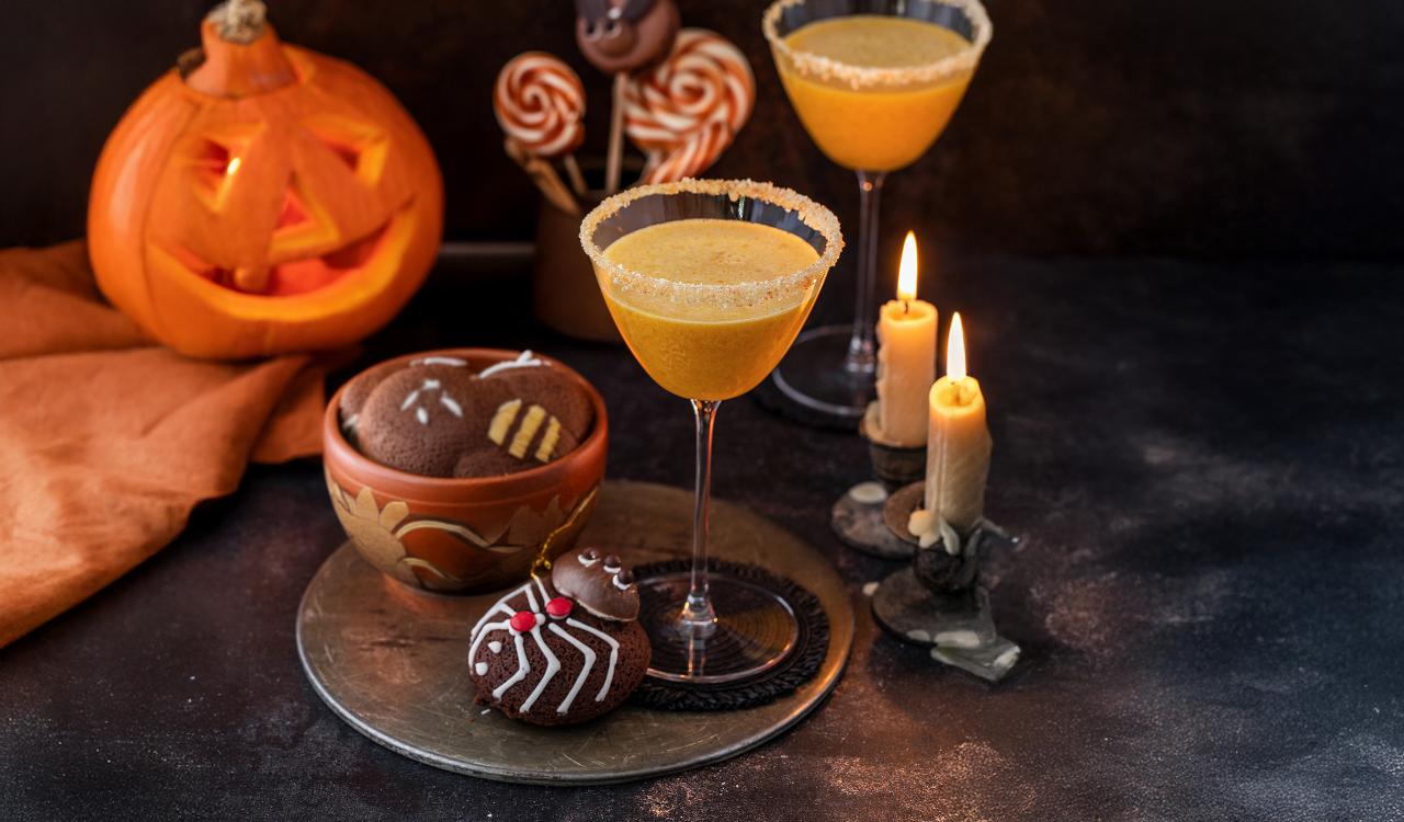 Zwei mit Zuckerrand verzierte Cocktailgläser mit orangefarbenem Getränk, daneben Halloween-Kürbis, Kerzen und Schokoladenkekse mit Spinnennetz-Dekor