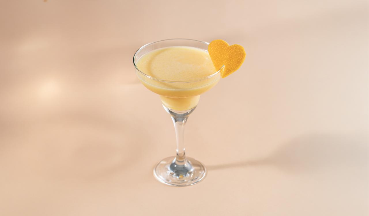Cocktailglas mit hellgelbem Getränk und herzförmiger Orangenschale am Glasrand vor beigem Hintergrund