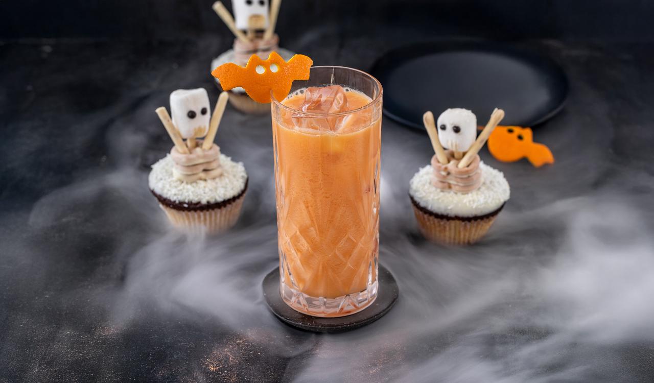 Glas mit orangefarbenem Getränk und Eiswürfeln auf schwarzem Untergrund umgeben von drei Cupcakes mit Skelett- und Fledermaus-Dekorationen und Nebeleffekt