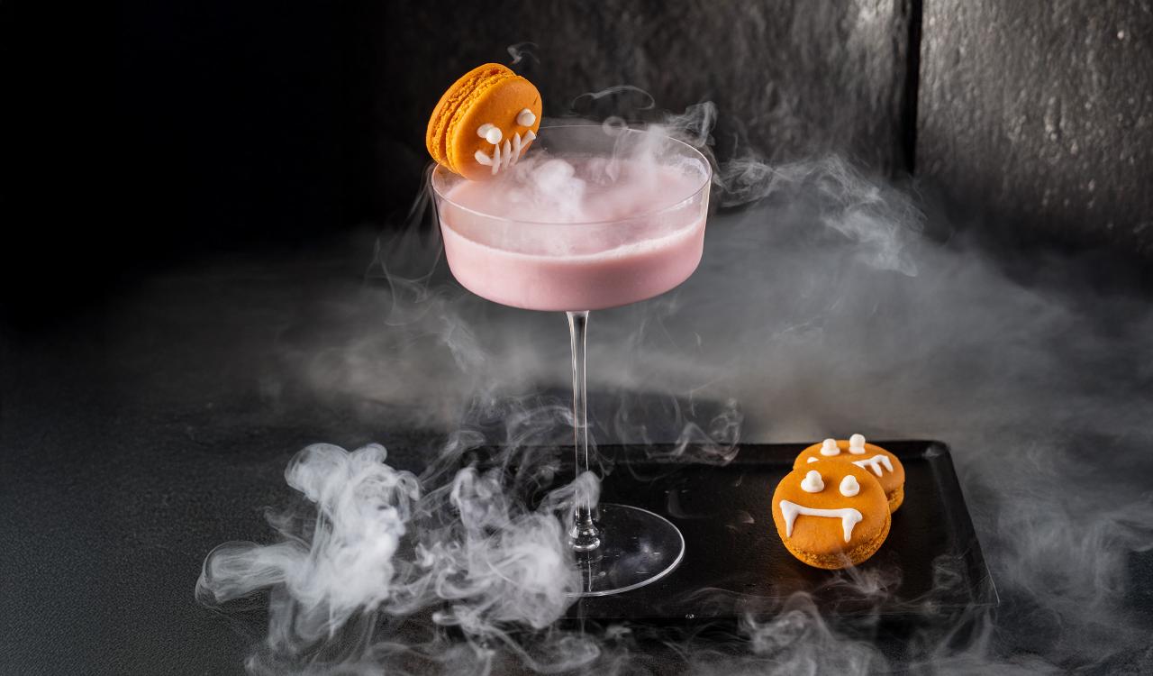 Cocktailglas mit rosa Getränk und orangefarbenem Keks mit Vampirzähnen auf Glasrand und schwarzem Tablett mit Nebeleffekt