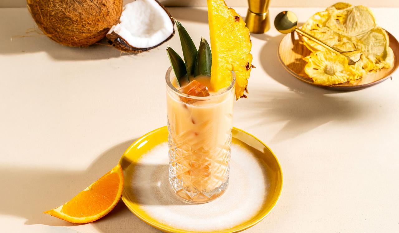 Glas mit hellbraunem Cocktail, garniert mit Ananasstück und grünen Blättern, auf weißem Teller mit gelbem Rand, daneben Orangenscheibe und halbierte Kokosnuss sowie goldfarbene Schale mit Eiswürfeln