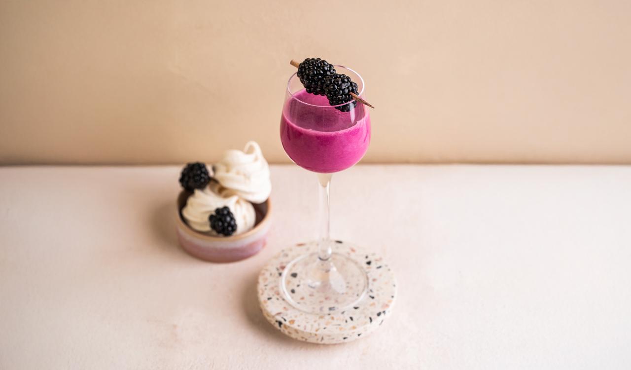 Glas mit pinkem Getränk und Brombeeren auf einem runden Steinuntersetzer neben zwei kleinen weißen Desserts mit Brombeeren auf hellem Untergrund