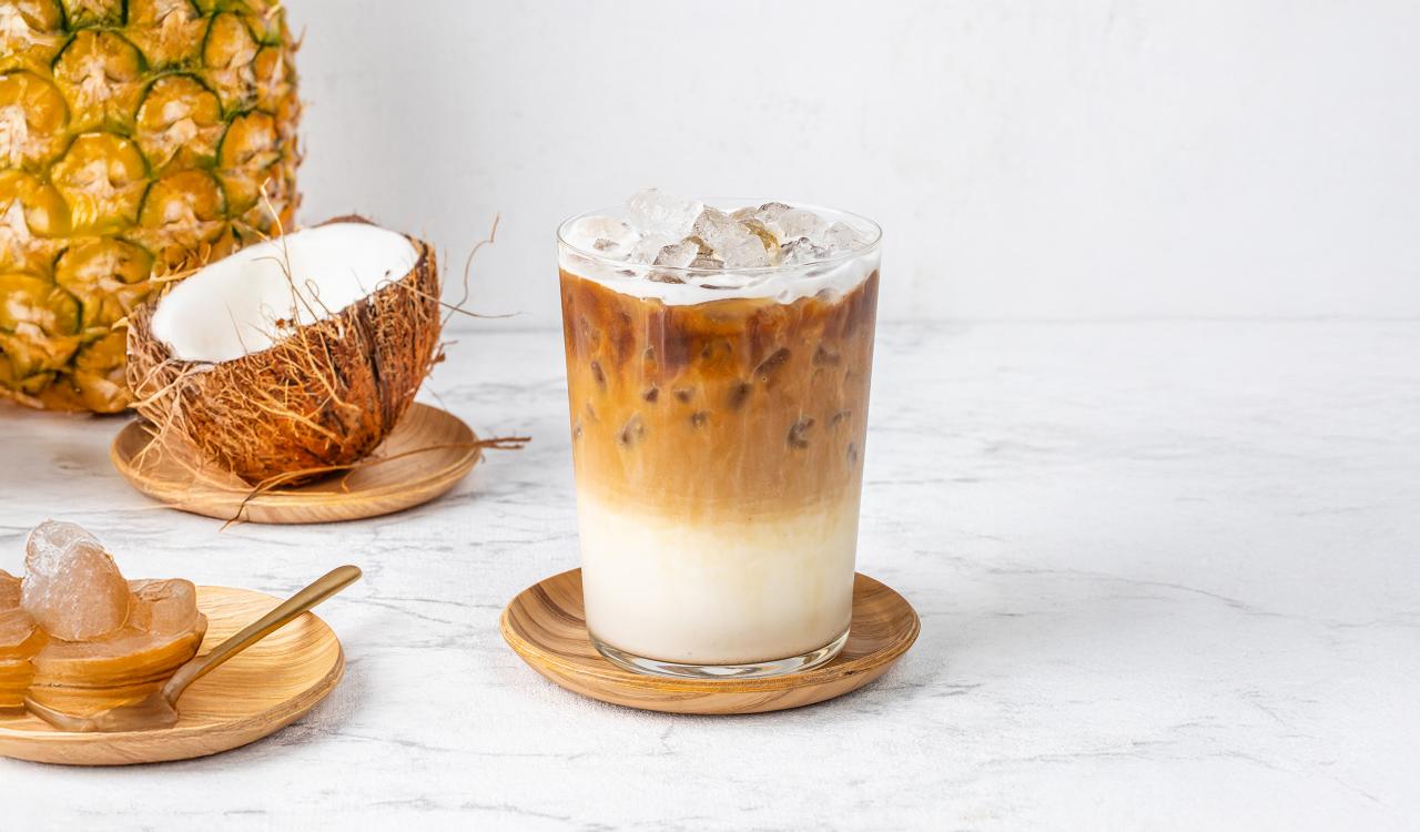 Glas mit Eiskaffee auf Holzuntersetzer vor halbierter Kokosnuss und Ananas auf weißer Marmoroberfläche