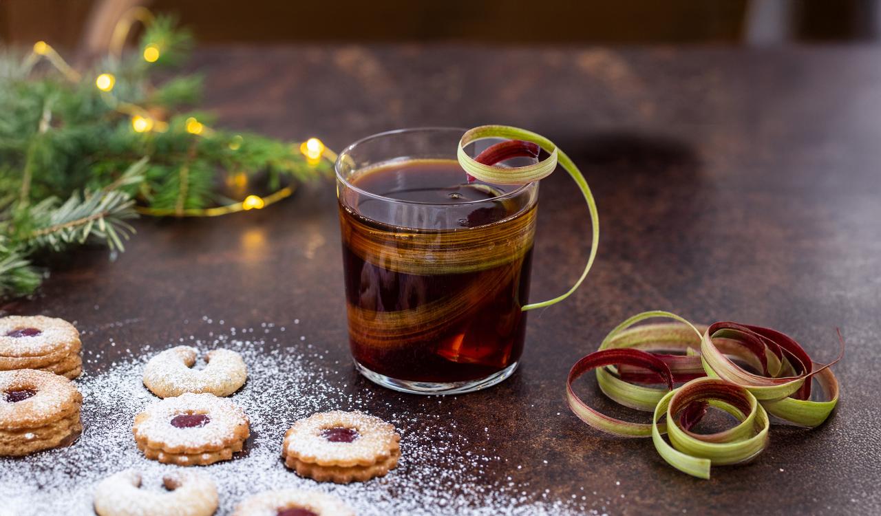 Glas mit dunklem Getränk und einer Spirale aus grün-rotem Gemüse oder Obst als Garnitur neben mehreren mit Puderzucker bestäubten Linzer Plätzchen auf dunklem Untergrund