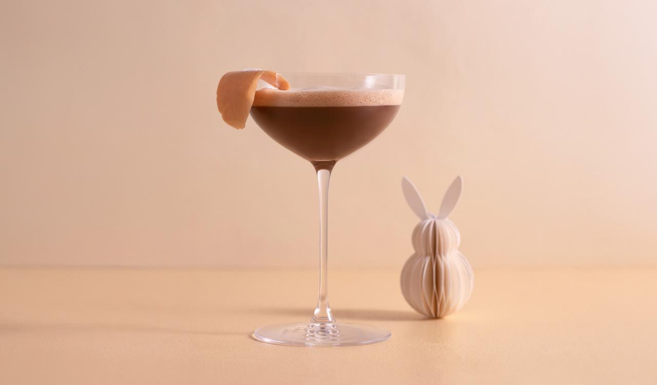 Cocktailglas mit braunem Getränk und Orangenzeste auf hellem Untergrund neben einer kleinen Papierfigur in Hasenform