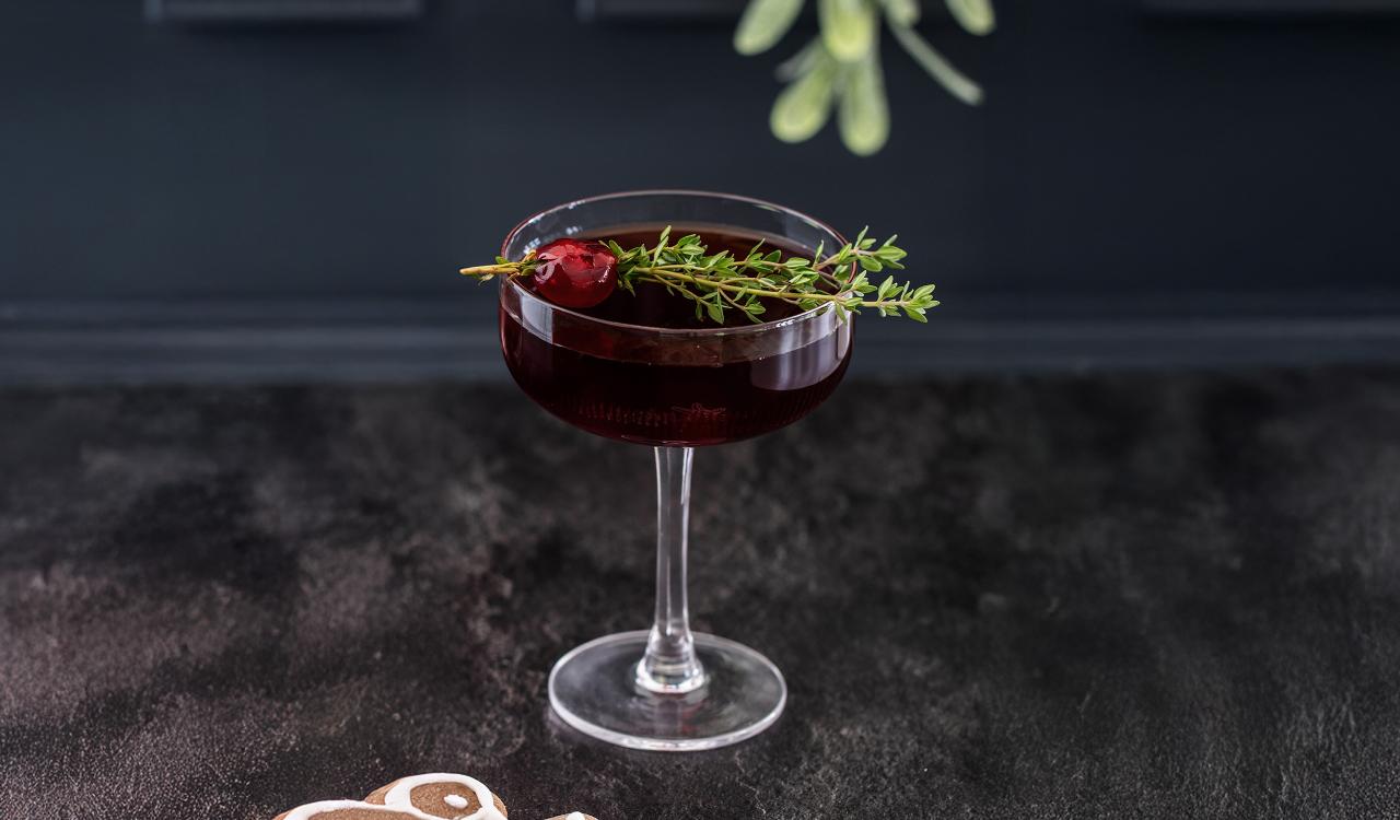 Cocktailglas mit dunklem Getränk, garniert mit einem Zweig Thymian und einer roten Beere